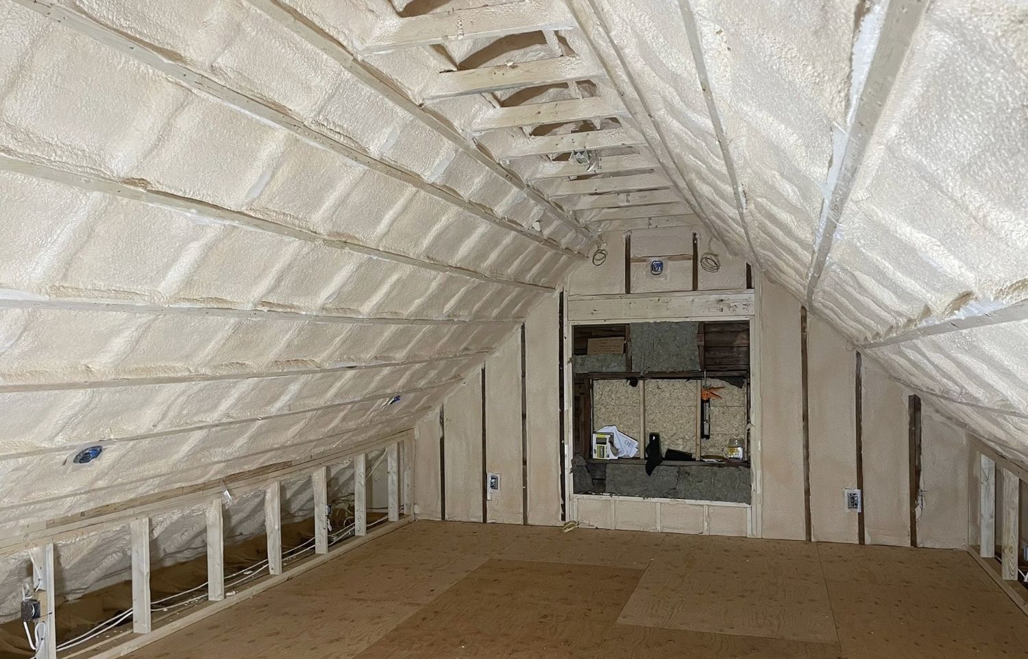 Attic room with both roof slopes and end wall fully covered in spray foam, plywood floor below