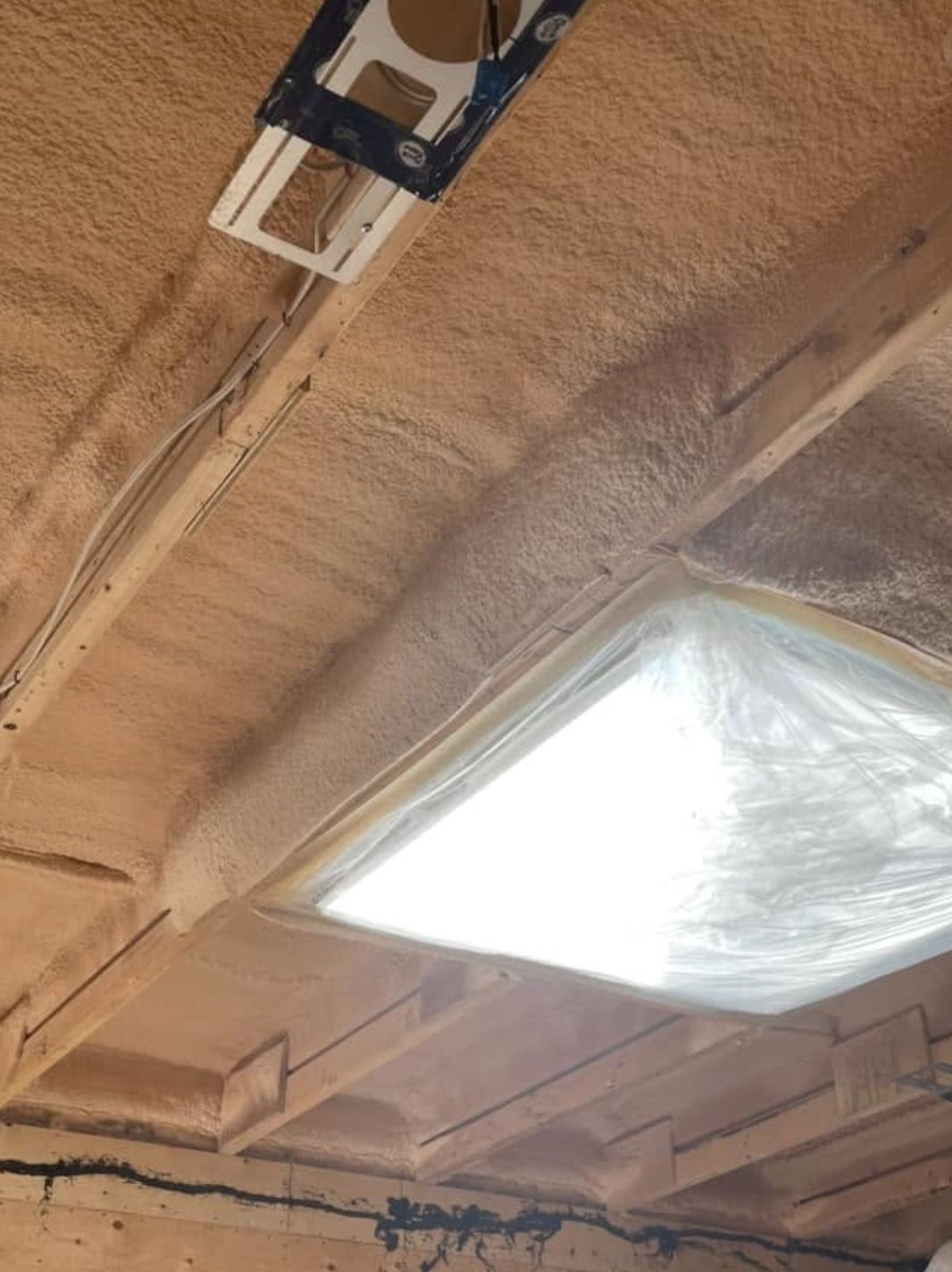 Spray foamed ceiling with a recessed light frame and a poly-covered opening letting in daylight