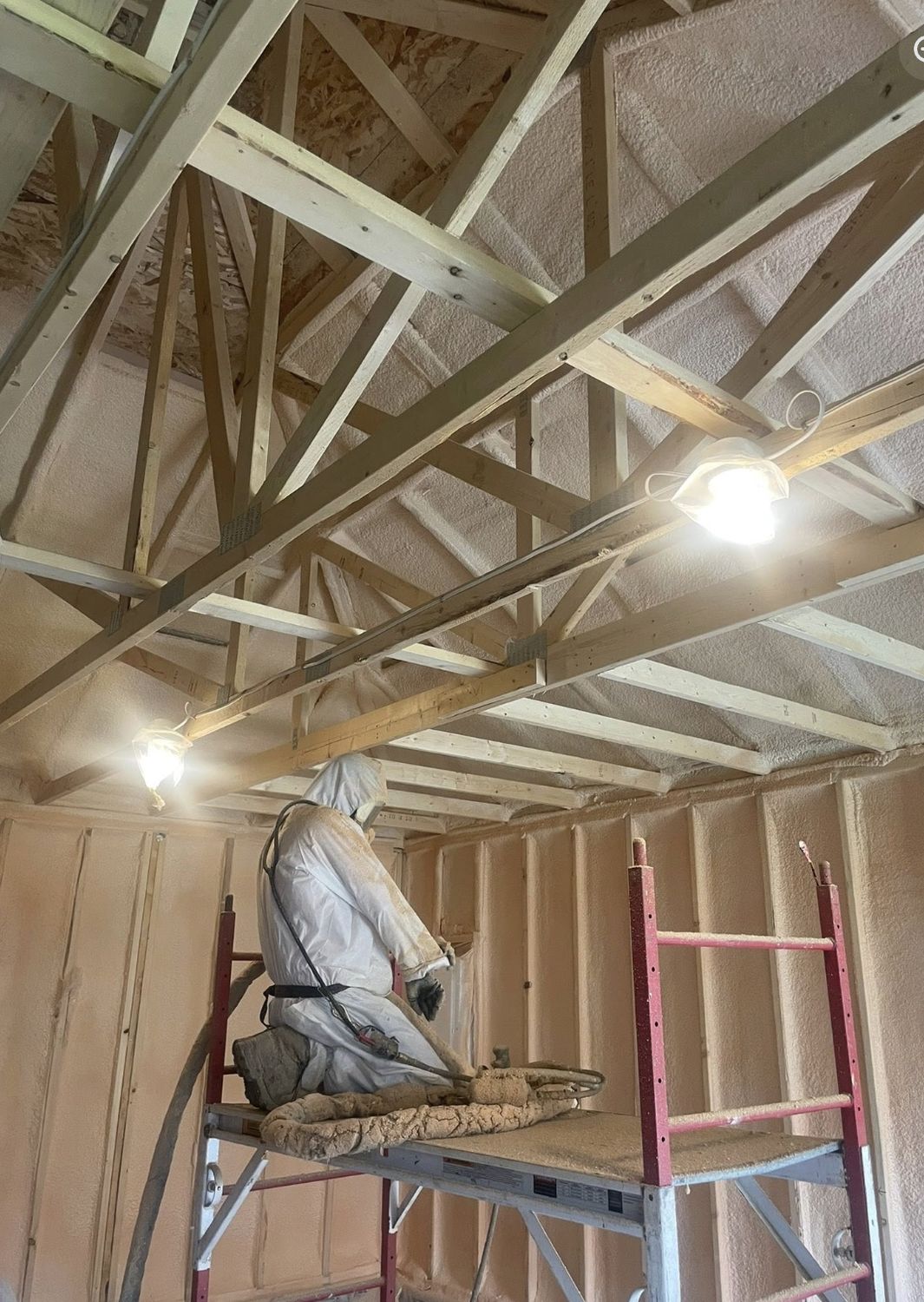 Installer in protective gear kneeling on a scaffold beneath roof trusses in a garage with spray foamed walls
