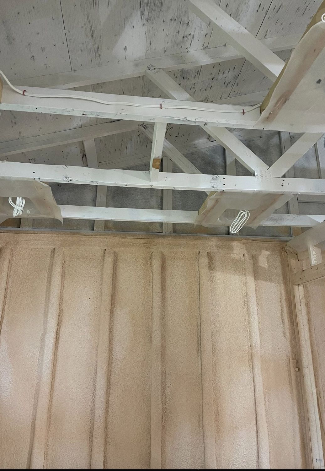 Spray foamed wall between wood strapping inside a garage with painted roof trusses above