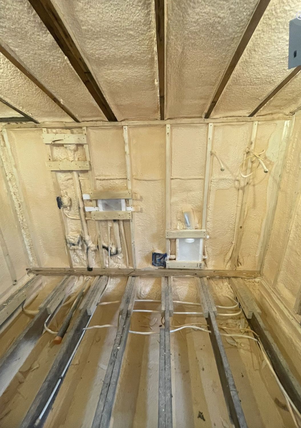 Small renovated room with spray foam covering walls and ceiling, plumbing rough-ins visible