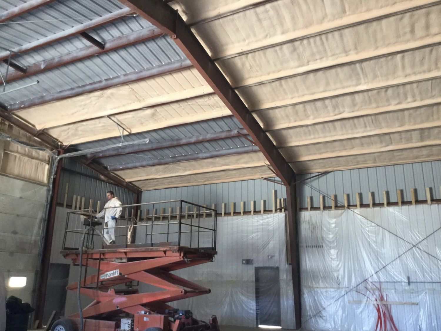 Installer on a scissor lift spray foaming the roof of a steel building between purlins